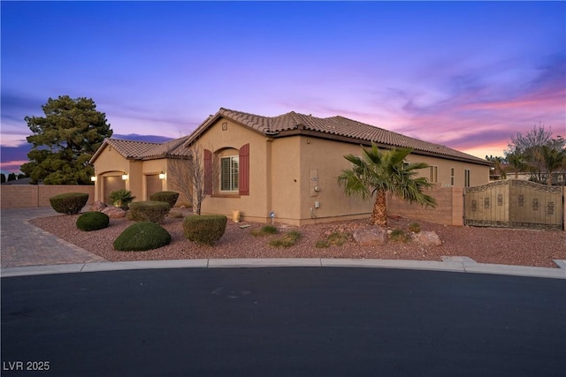 property exterior at dusk featuring decorative driveway, a tile roof, stucco siding, an attached garage, and fence