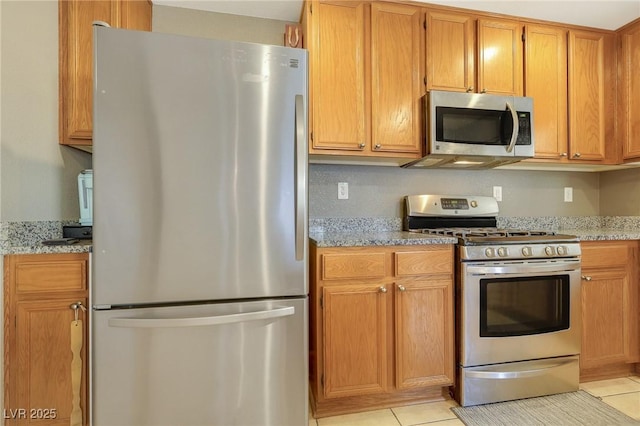 kitchen featuring light tile patterned floors, appliances with stainless steel finishes, brown cabinets, and light stone counters