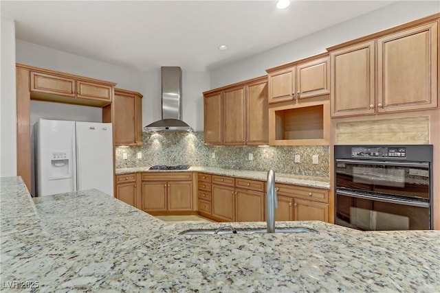 kitchen with light stone counters, dobule oven black, stainless steel gas stovetop, wall chimney range hood, and white fridge with ice dispenser