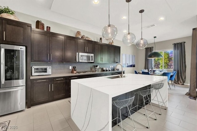 kitchen featuring appliances with stainless steel finishes, decorative light fixtures, a sink, a kitchen island with sink, and backsplash