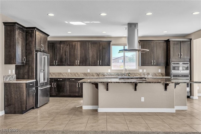 kitchen with stainless steel appliances, dark stone countertops, dark brown cabinets, island range hood, and a kitchen breakfast bar