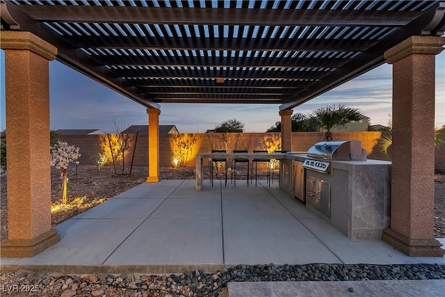 patio terrace at dusk featuring exterior kitchen, grilling area, a fenced backyard, and a pergola