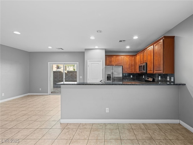 kitchen with tasteful backsplash, appliances with stainless steel finishes, brown cabinetry, dark stone counters, and a peninsula