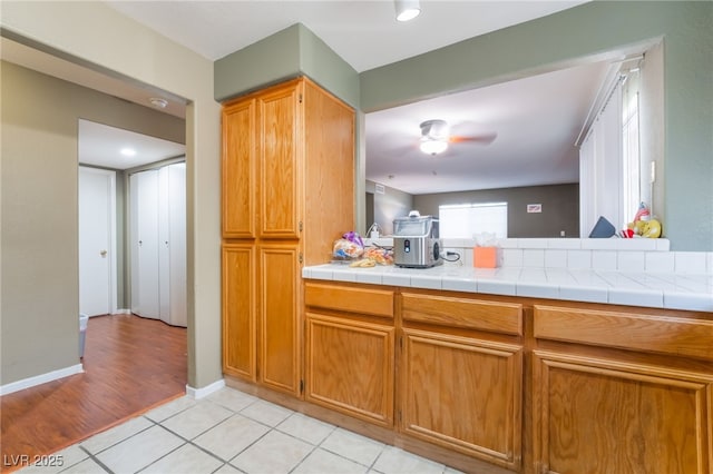 kitchen featuring light tile patterned floors, ceiling fan, baseboards, tile counters, and brown cabinetry