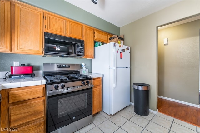 kitchen featuring tile countertops, freestanding refrigerator, stainless steel gas stove, light tile patterned flooring, and black microwave