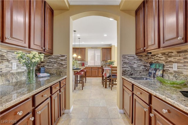 kitchen featuring arched walkways, brown cabinetry, decorative light fixtures, light stone countertops, and a sink