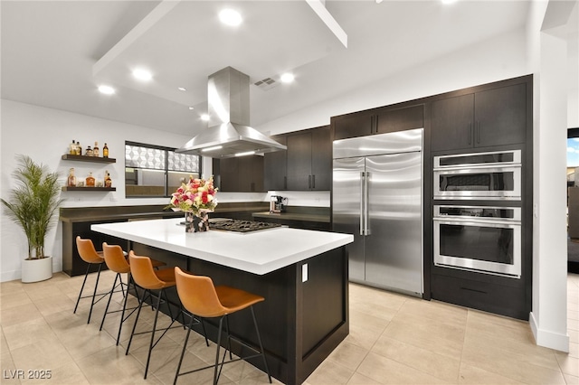 kitchen featuring island exhaust hood, open shelves, appliances with stainless steel finishes, a kitchen island, and a kitchen bar