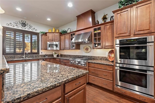 kitchen featuring recessed lighting, wood finished floors, appliances with stainless steel finishes, dark stone counters, and wall chimney exhaust hood