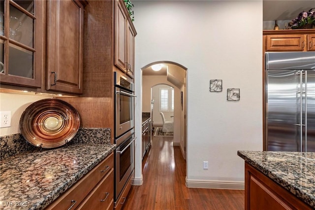 kitchen with arched walkways, dark wood finished floors, glass insert cabinets, dark stone countertops, and stainless steel appliances