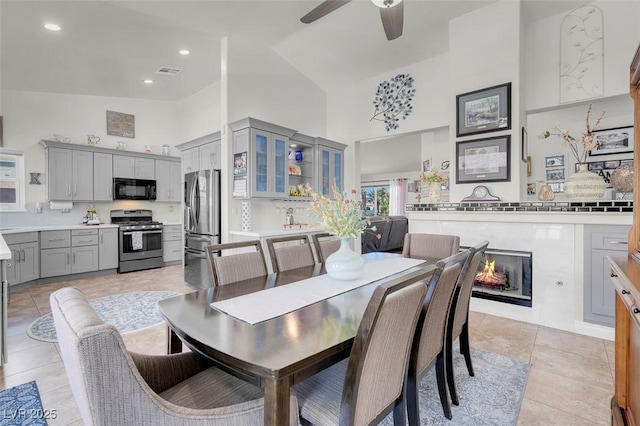 dining room with a warm lit fireplace, light tile patterned floors, a high ceiling, visible vents, and a ceiling fan