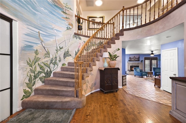 stairway with a fireplace, visible vents, a towering ceiling, a ceiling fan, and wood finished floors