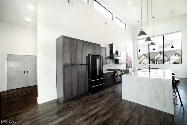 kitchen featuring premium appliances, an island with sink, dark wood-style floors, hanging light fixtures, and wall chimney range hood