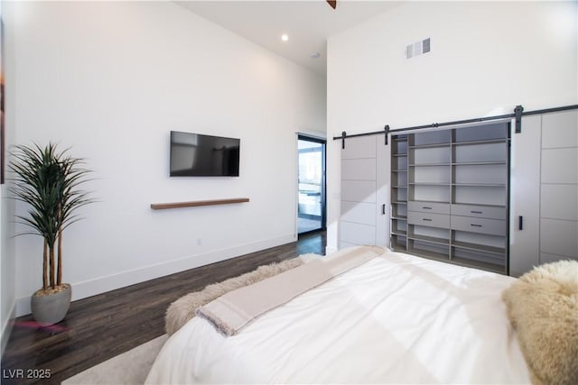 bedroom featuring a barn door, a high ceiling, visible vents, baseboards, and dark wood finished floors