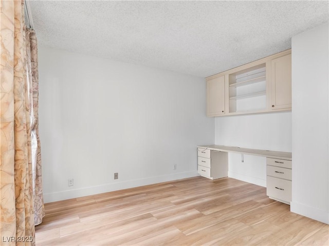 unfurnished office with a textured ceiling, built in desk, light wood-type flooring, and baseboards