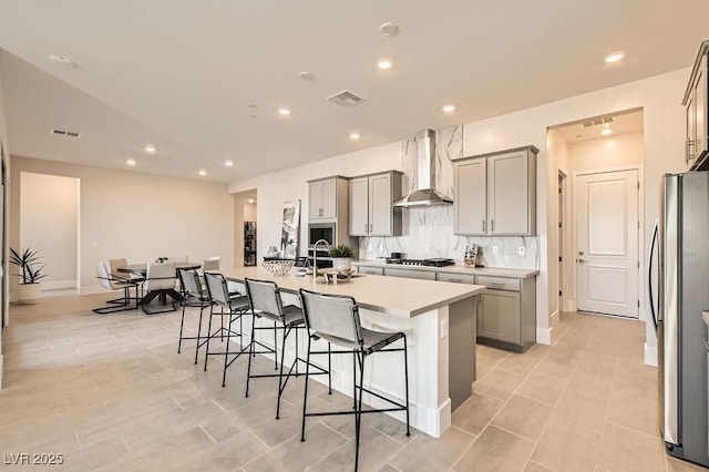 kitchen with an island with sink, wall chimney range hood, appliances with stainless steel finishes, and gray cabinetry