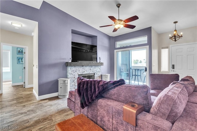 living area featuring baseboards, lofted ceiling, light wood-style flooring, a stone fireplace, and ceiling fan with notable chandelier