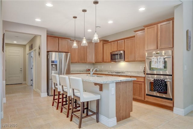 kitchen with a kitchen island with sink, stainless steel appliances, a sink, light stone countertops, and pendant lighting