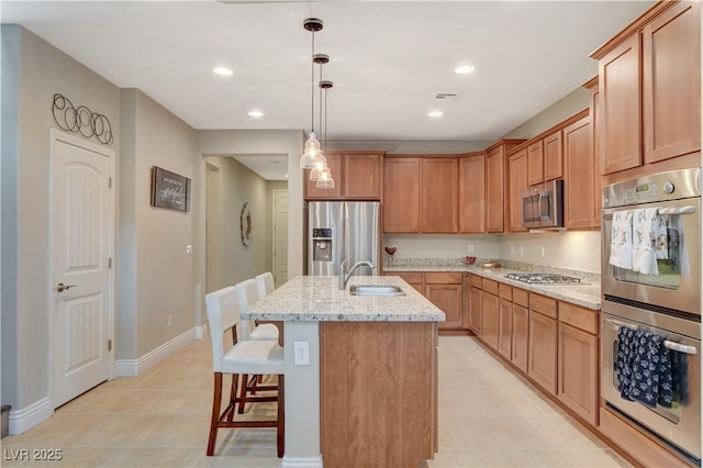 kitchen with a center island with sink, appliances with stainless steel finishes, light stone counters, hanging light fixtures, and a sink