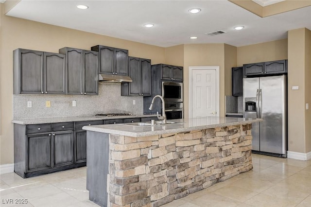 kitchen with under cabinet range hood, a sink, light countertops, appliances with stainless steel finishes, and a center island with sink