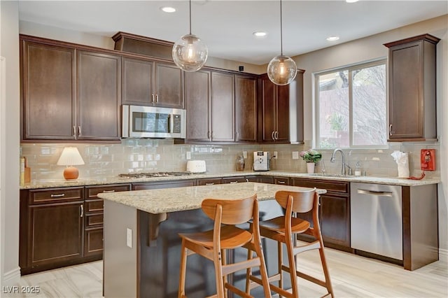 kitchen featuring stainless steel appliances, a kitchen island, a sink, light stone countertops, and pendant lighting