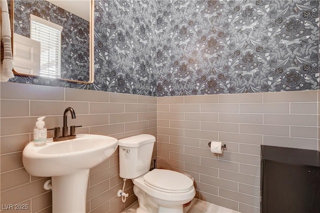 half bathroom with toilet, wallpapered walls, tile walls, and wainscoting