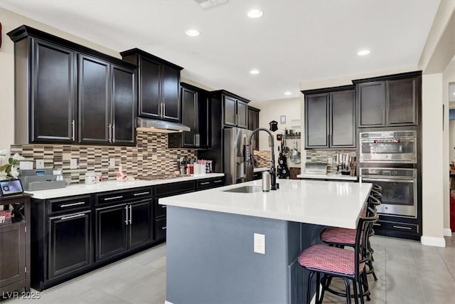 kitchen with appliances with stainless steel finishes, dark cabinets, under cabinet range hood, a kitchen bar, and a sink