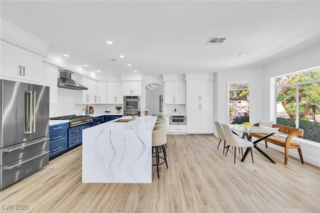 kitchen with blue cabinets, white cabinetry, visible vents, appliances with stainless steel finishes, and wall chimney range hood