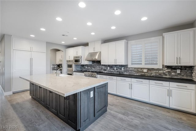 kitchen featuring arched walkways, light wood-style floors, white cabinets, a kitchen island with sink, and under cabinet range hood