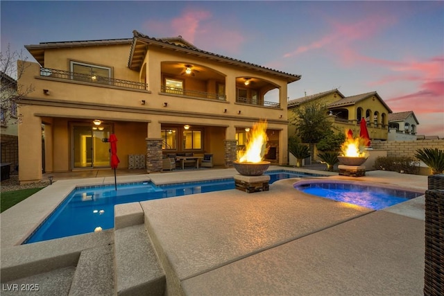 back of property at dusk featuring a fenced in pool, a patio, an outdoor fire pit, a balcony, and an in ground hot tub