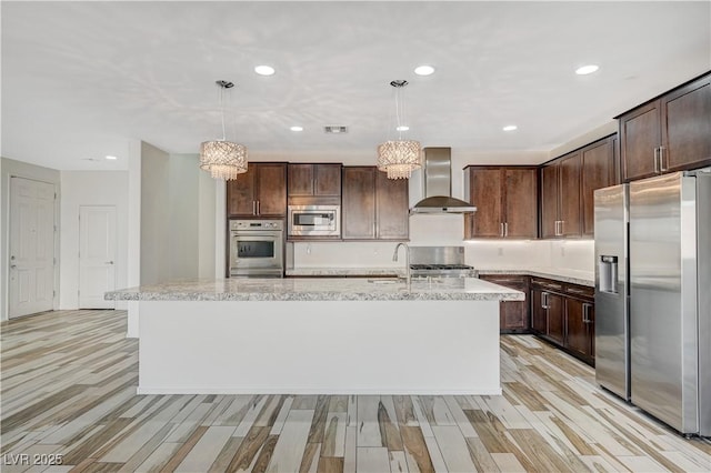 kitchen with a center island with sink, light wood-style flooring, appliances with stainless steel finishes, dark brown cabinets, and wall chimney range hood