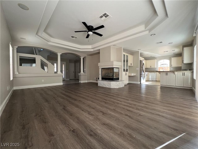 unfurnished living room with arched walkways, a tray ceiling, ornate columns, visible vents, and a multi sided fireplace