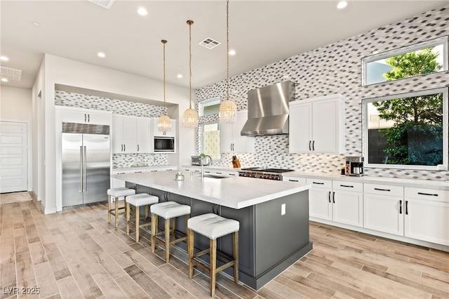 kitchen featuring visible vents, built in appliances, a kitchen island with sink, light countertops, and wall chimney range hood