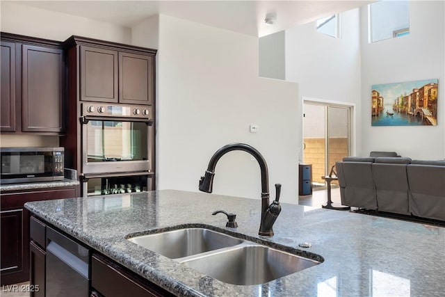 kitchen with dark brown cabinetry, stainless steel appliances, a sink, open floor plan, and dark stone countertops