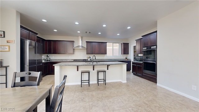 kitchen with dobule oven black, an island with sink, wall chimney exhaust hood, a kitchen breakfast bar, and freestanding refrigerator