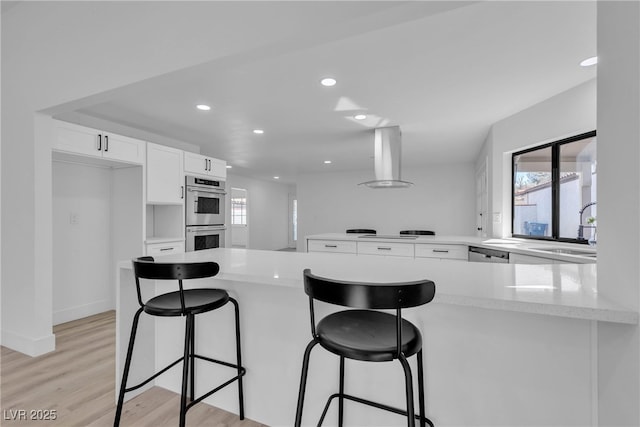 kitchen with a peninsula, wall chimney range hood, double oven, white cabinetry, and a sink