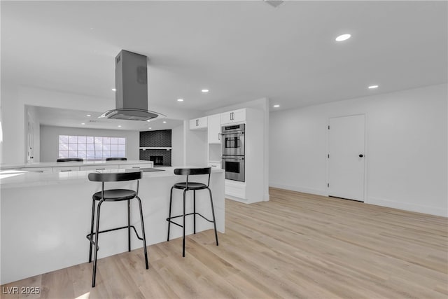 kitchen featuring island exhaust hood, light countertops, light wood-style floors, stainless steel double oven, and white cabinetry