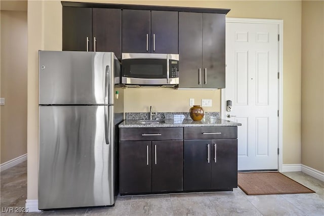 kitchen featuring light stone counters, stainless steel appliances, dark brown cabinetry, a sink, and baseboards