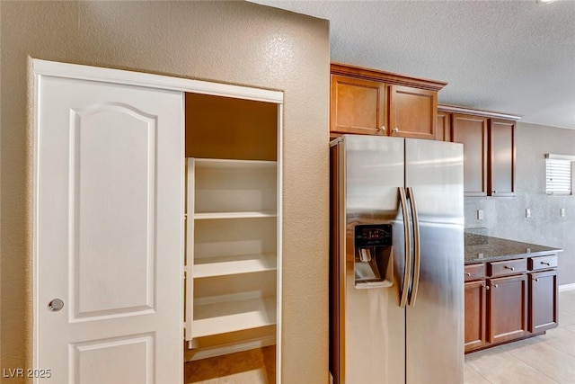 kitchen featuring light tile patterned floors, brown cabinetry, a textured ceiling, dark stone counters, and stainless steel fridge