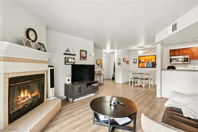 living room featuring light wood-type flooring, a glass covered fireplace, visible vents, and ceiling fan