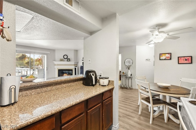 kitchen with a textured ceiling, ceiling fan, light wood-style flooring, visible vents, and a glass covered fireplace