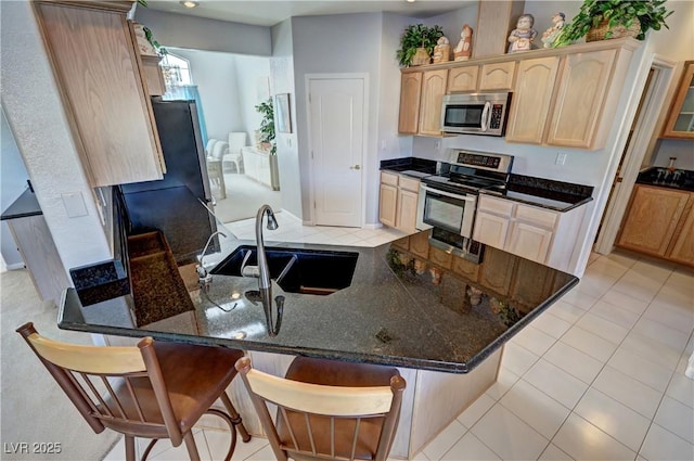 kitchen featuring dark stone counters, a peninsula, stainless steel appliances, light brown cabinets, and a sink