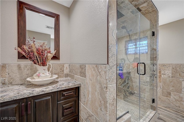 full bath featuring a stall shower, visible vents, a textured wall, vanity, and tile walls