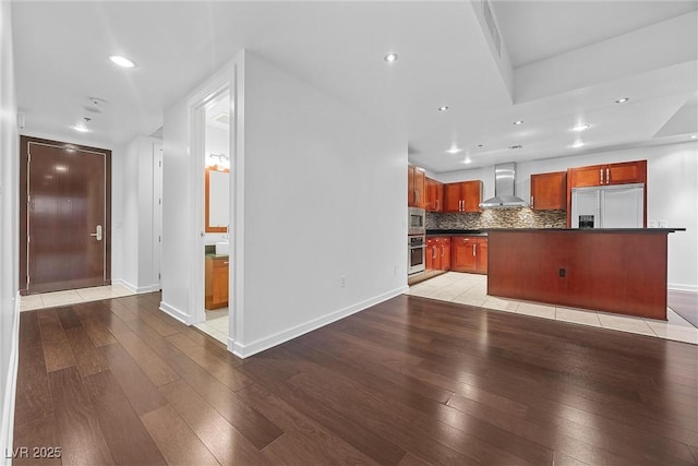 kitchen with dark countertops, backsplash, built in appliances, light wood-type flooring, and wall chimney exhaust hood