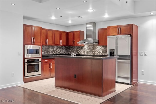 kitchen with built in appliances, wall chimney range hood, dark countertops, and decorative backsplash