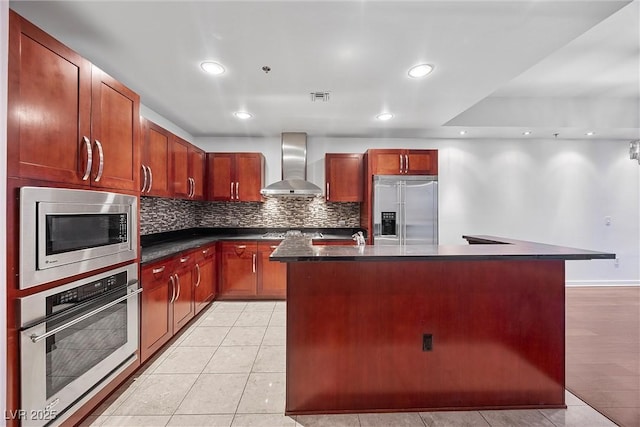 kitchen featuring built in appliances, backsplash, wall chimney exhaust hood, dark countertops, and a center island with sink