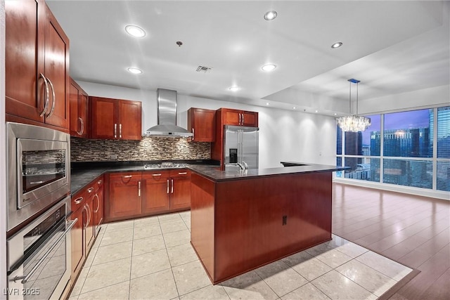 kitchen with dark countertops, visible vents, a kitchen island with sink, built in appliances, and wall chimney exhaust hood