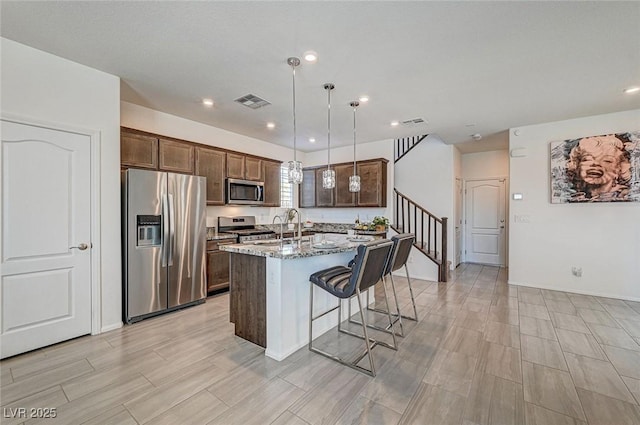 kitchen featuring a breakfast bar area, a kitchen island with sink, appliances with stainless steel finishes, light stone countertops, and pendant lighting