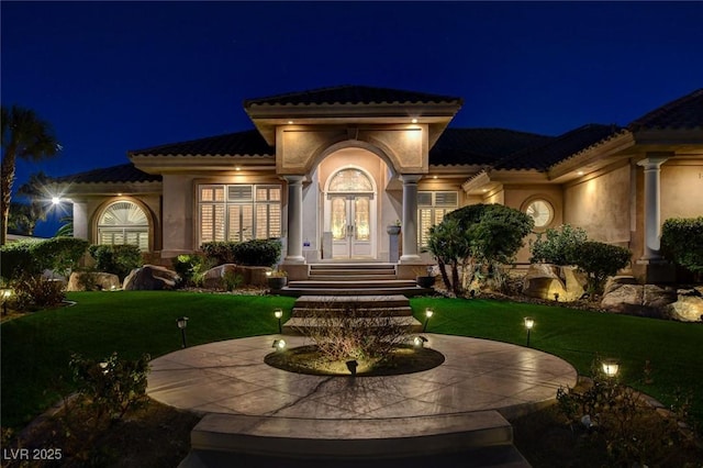 exterior entry at night featuring a tiled roof, french doors, a lawn, and stucco siding