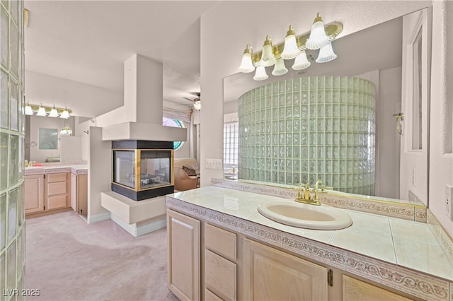 bathroom featuring a ceiling fan, two vanities, a sink, and a multi sided fireplace