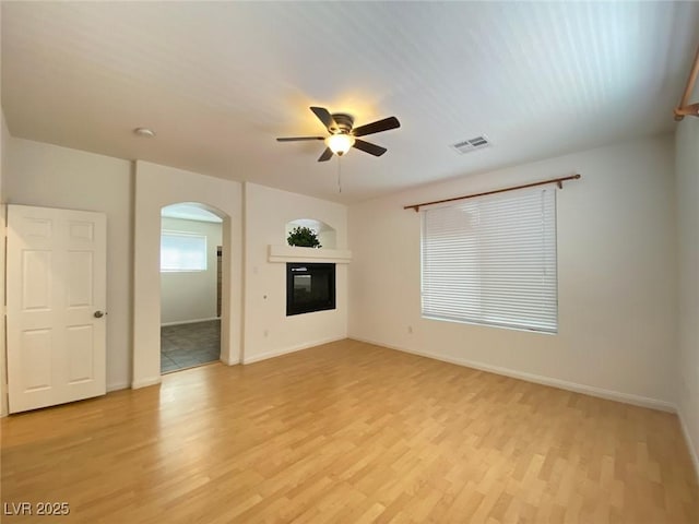 unfurnished living room featuring arched walkways, light wood-style flooring, visible vents, a ceiling fan, and a glass covered fireplace